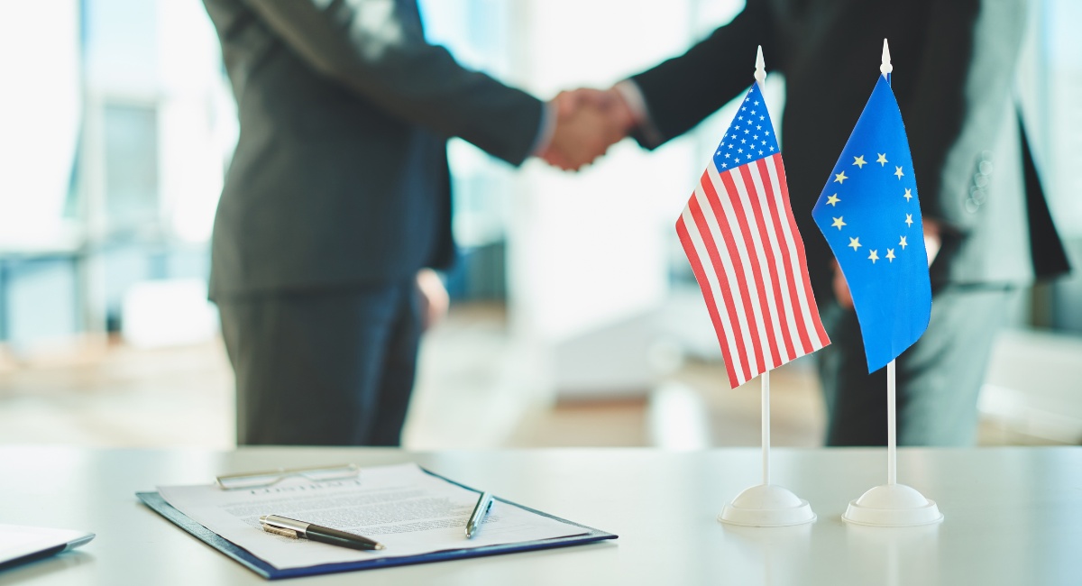 Two hands clasping behind the American and European flags, highlighting the agreement signed for the tariffs imposed by the USA on Europe.