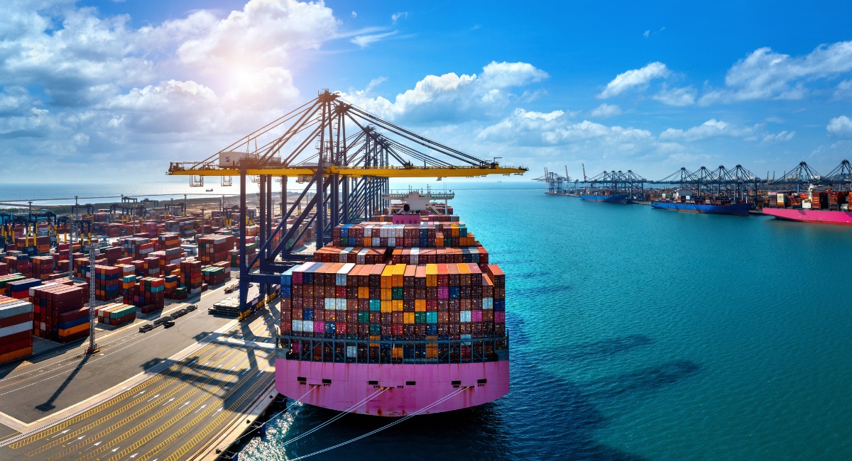 Nave cargo piena di container, attraccata presso un porto industriale, sta venendo scaricata con delle grandi gru