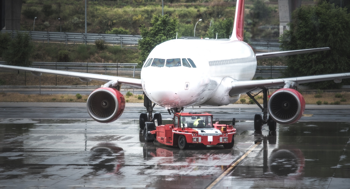Aereo passeggeri trainato da un trattore aeroportuale su pista bagnata, per simboleggiare la carenza di carburante aereo in Europa nel 2026.